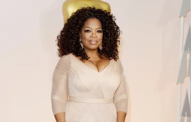 A person in a formal beige dress stands in front of a golden Oscar statue.