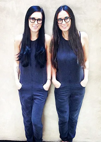 Two women with long dark hair and glasses, wearing dark jumpsuits, standing side by side against a plain background.