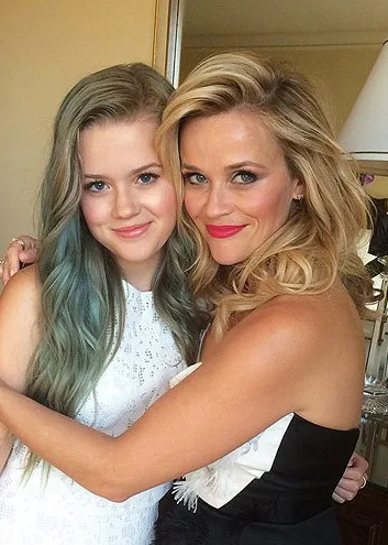 Two women smiling, embracing each other warmly. One has long blue-tinted hair, the other has curly blonde hair.