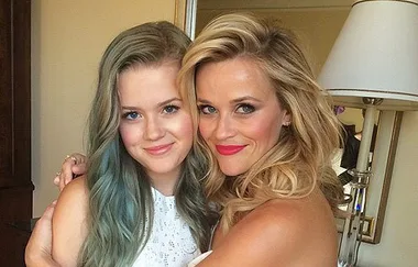 Two women smiling and embracing, one with long blonde hair and the other with long blue-tinted hair, indoors.
