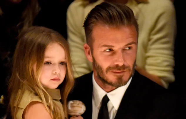 A man in a formal suit holding a young girl with long hair, both looking to the side in a dimly lit setting.