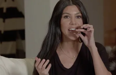 A person with long dark hair eating something while seated on a couch, wearing a black top indoors.