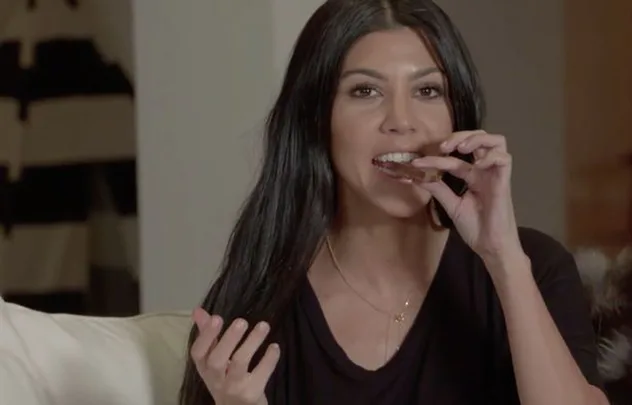 A person with long dark hair eating something while seated on a couch, wearing a black top indoors.