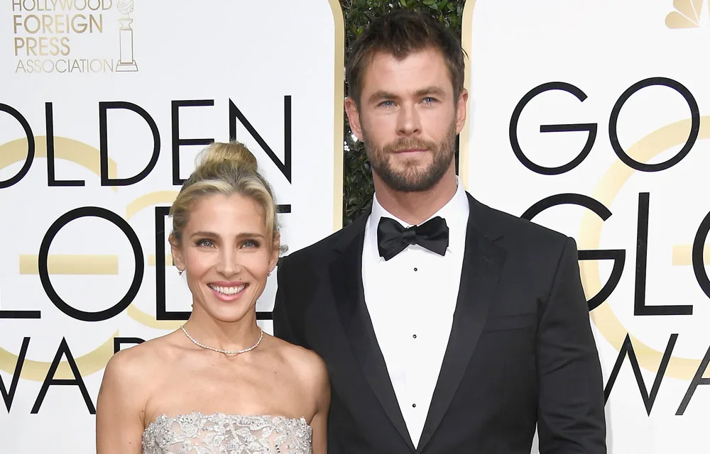 A man in a tuxedo and a woman in a strapless dress pose at the Golden Globe Awards with a backdrop featuring the event's logo.
