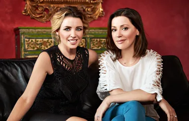 Two women sitting on a black sofa against a red wall with ornate decor, smiling at the camera.