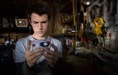 A young man holds and examines a cassette tape in a dimly lit workshop.