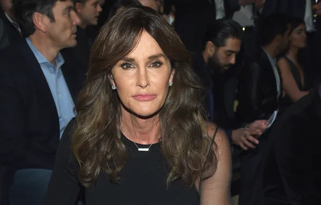 Person with long wavy hair in a black outfit sits in a crowded event setting, looking toward the camera.