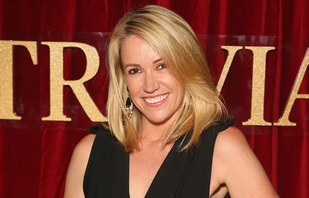 A smiling person with blonde hair stands against a red background with gold lettering, wearing a black outfit.