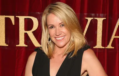 A smiling person with blonde hair stands against a red background with gold lettering, wearing a black outfit.