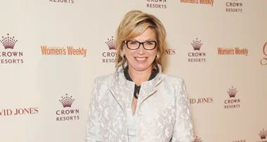 A woman in glasses and a patterned white jacket smiles at an event with logos in the background.