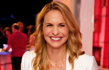 Smiling woman with long blonde hair in a white jacket, standing in a room with people and warm lighting.