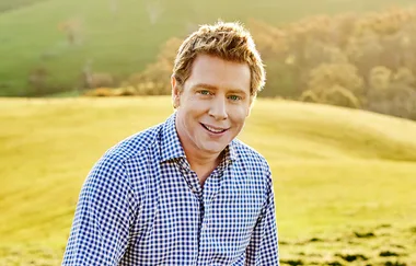 Man in a checkered shirt smiles in a sunny green field landscape.