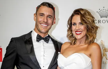 Couple in formal attire smiling at an event, man in a black tuxedo and woman in a white strapless dress.
