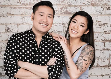 Two people smiling, one in a polka dot shirt, the other with arm tattoos, standing against a brick wall.