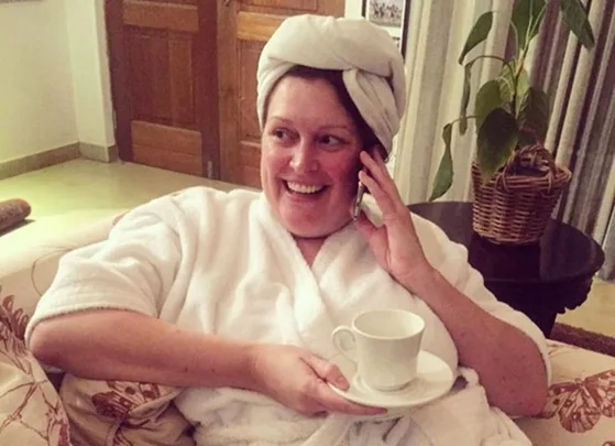 Person in a white robe and towel on head, smiling, holding a cup, seated indoors.