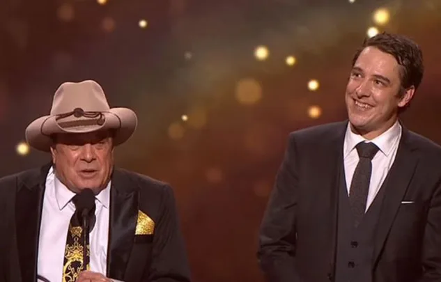 Two men on stage, one wearing a cowboy hat, both in suits, smiling under warm spotlights.