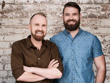 Two smiling men standing in front of a textured brick wall, one in a brown shirt and the other in a blue patterned shirt.
