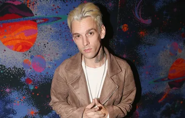 Man with blonde hair and a light brown jacket stands in front of a colorful space-themed background.