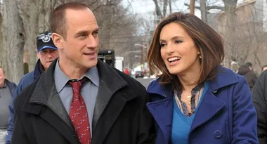 Man in a suit and woman in a blue coat walking outdoors, both smiling, with people and trees in the background.