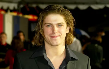 Young man with long hair wearing a dark collared shirt and blazer smiles at an event, with a blurred background.