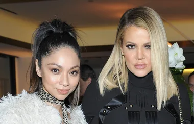 Two women posing at an event, one with a high ponytail and faux fur coat, the other with blonde hair and a black outfit.