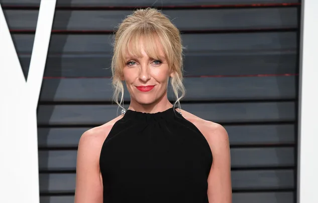 Woman with blonde hair in a black halter dress, posing indoors against a striped background.