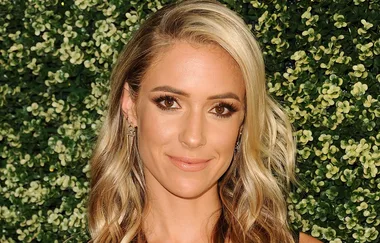 Blonde woman with wavy hair in front of a leafy green background, smiling slightly.