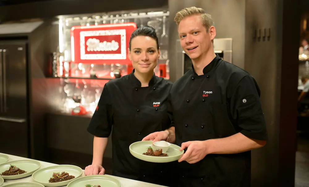 Two chefs in black uniforms presenting a dish in a kitchen setting.