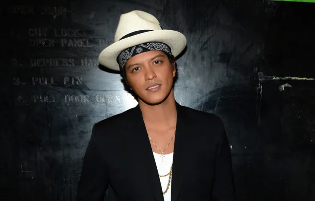 Singer in a black jacket, white shirt, bandana, straw fedora, and gold chain against a dark backdrop.