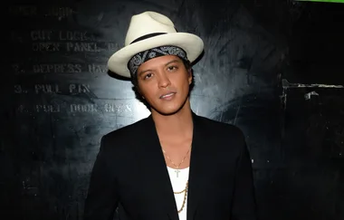 Singer in a black jacket, white shirt, bandana, straw fedora, and gold chain against a dark backdrop.