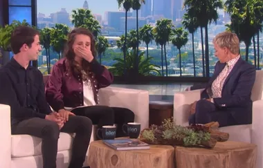 Two guests on a talk show sit on a couch, one looks surprised, while the host listens.