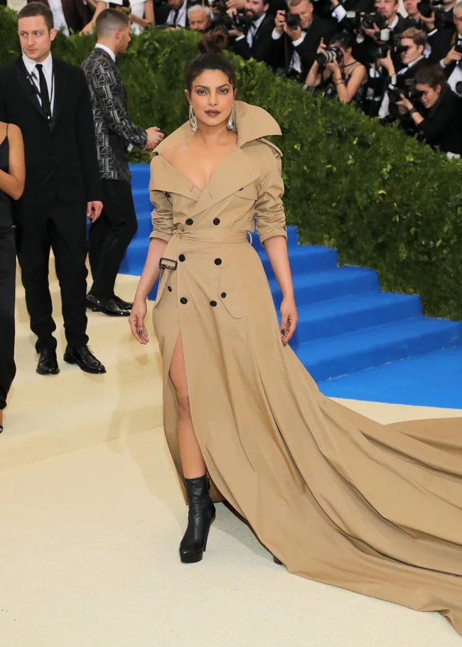 Person in a tan trench coat gown, black boots at a formal event with photographers and a blue carpeted stairway.