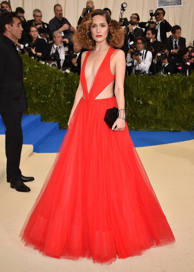 A woman in a vibrant red gown poses on a beige carpet at a formal event, surrounded by photographers.