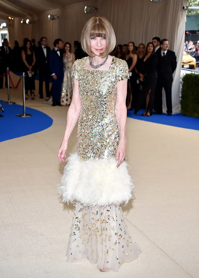 A woman in a sparkling dress with a feathered hem walks on a carpeted event floor, surrounded by an audience.