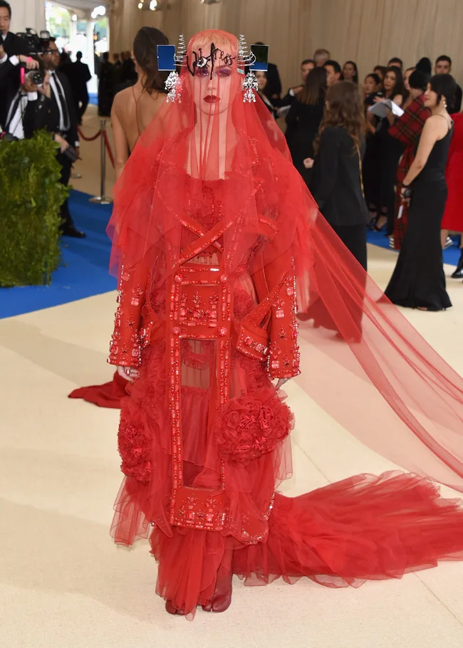 A person in an elaborate red gown with a veil and headpiece on a fashion event carpet.