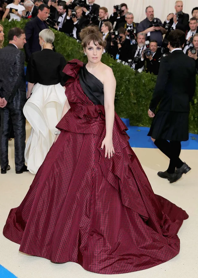 Woman in a red checkered gown with a ruffled design at a formal event with photographers in the background.