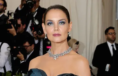A woman in a strapless gown and necklace poses on a red carpet, surrounded by photographers.