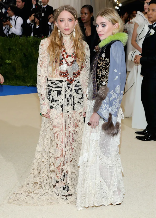 Two women in elaborate lace and embroidered dresses pose on a red carpet event, surrounded by onlookers.
