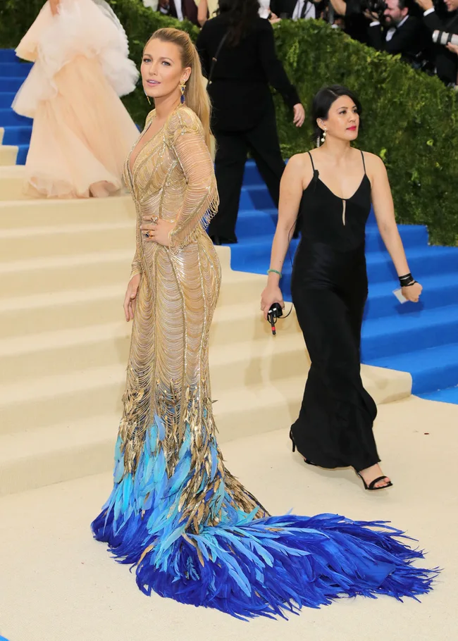 A woman in a gold and blue feathered gown on a carpeted staircase at a formal event.