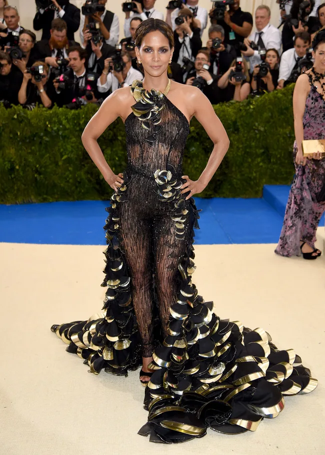 Woman in a dramatic black and gold gown poses on a red carpet, surrounded by photographers.