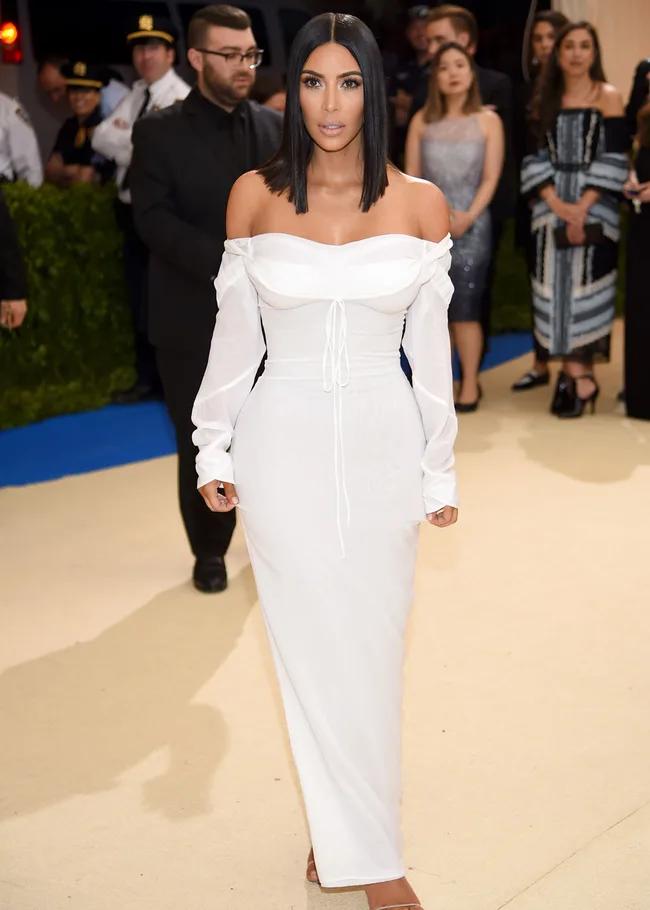 Woman in an elegant white off-shoulder gown at a formal event, with people in the background.