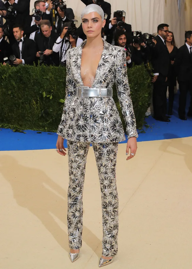 Person in silver embellished suit with white painted head at a formal event with photographers in the background.