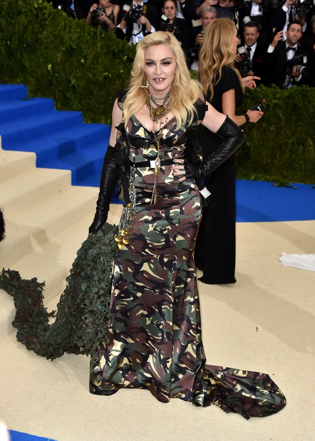 Woman in camo gown with long black gloves poses on a red carpet, photographers in the background.