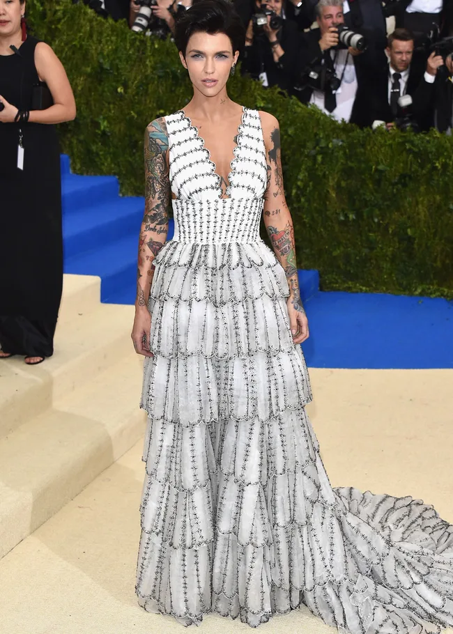 Individual in a layered white gown with tattoos, posing on a red carpet with photographers in the background.