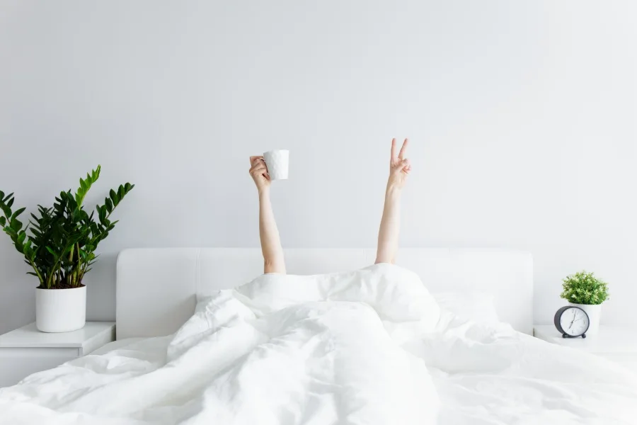 Woman under bed covers holding up a coffee