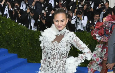 A person in a white lace dress poses on a blue carpet at an event, surrounded by photographers.