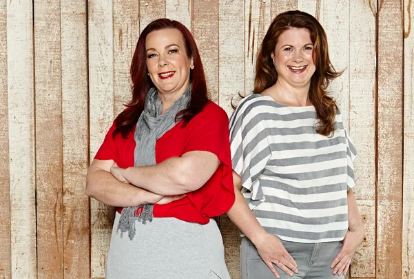 Two women smiling, one in a red top with a grey scarf and the other in a striped shirt, standing against a wooden background.