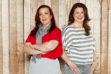 Two women smiling, one in a red top with a grey scarf and the other in a striped shirt, standing against a wooden background.