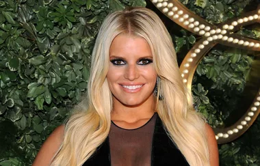 ```html
<img src="https://dankanator.com/wp-content/uploads/2020/01/jessica-simpson-in-her-new-book-opens-up-about-her-emotional-affair-with-johnny-knoxville-1.jpg" alt="Person with long blonde hair, wearing a black outfit, smiling in front of a green background." />
```