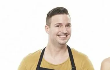 A man wearing a mustard shirt and a black apron, smiling against a white background.